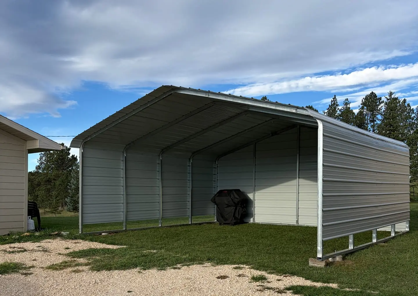 A covered metal carport with a charcoal grill, set on gravel beside the lawn with pines behind
