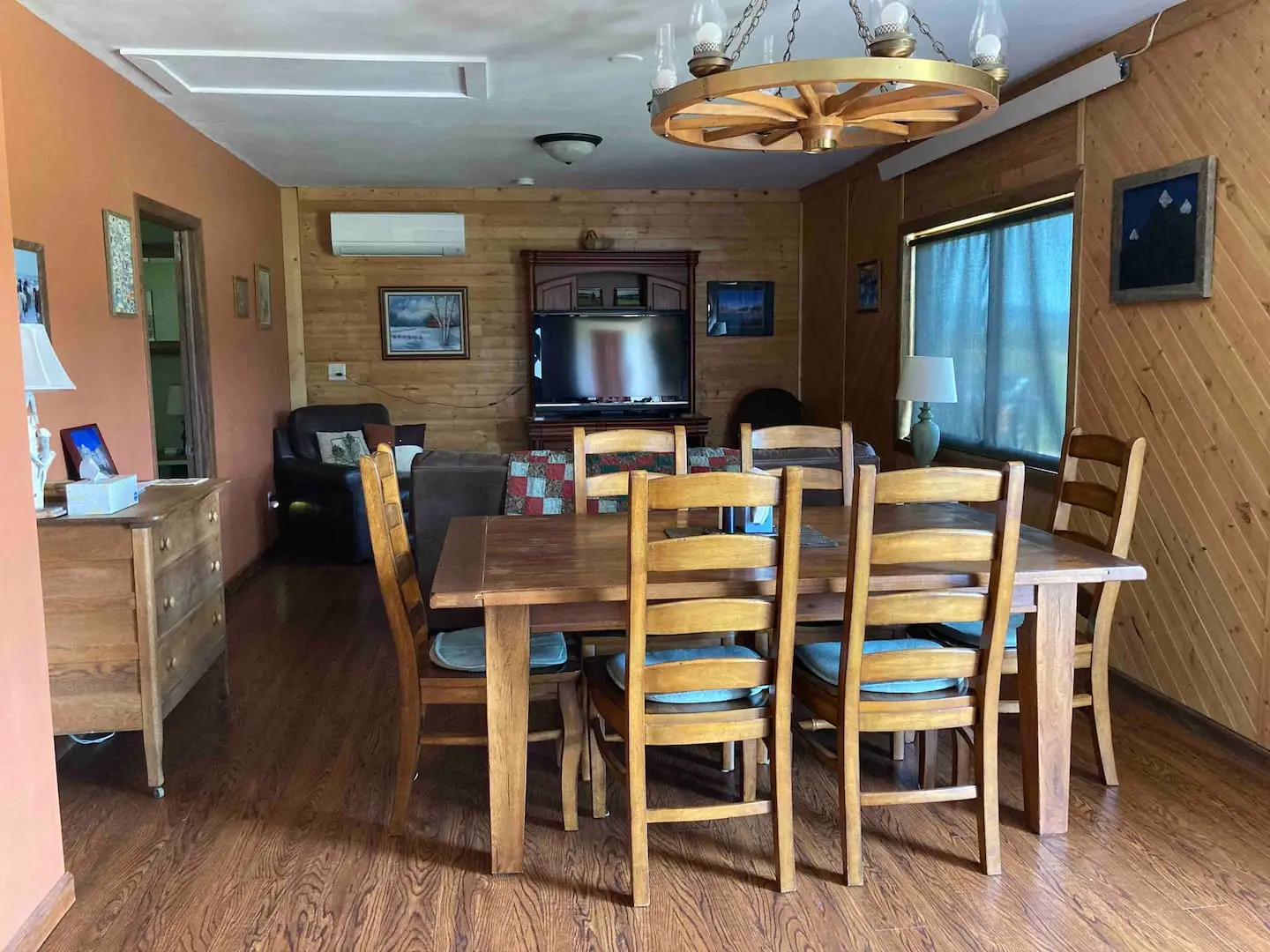 Open dining area with a long wooden dining table, a wagon-wheel chandelier and knotty-pine walls