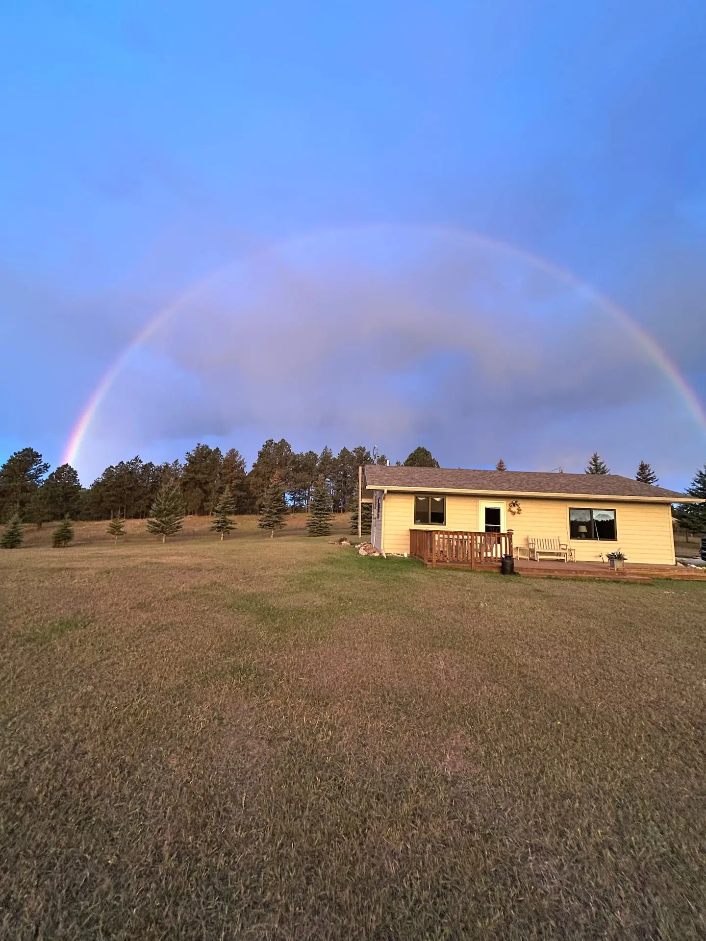 A full rainbow arcing over the cabin on its hilltop meadow, ringed by ponderosa pines