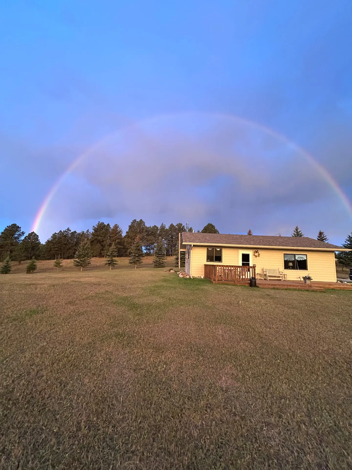 A full rainbow arcing over the cabin on its hilltop meadow, ringed by ponderosa pines